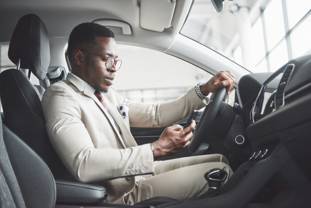 Stylish black businessman sitting behind the wheel of new luxury car. Rich african american man using smartphone.