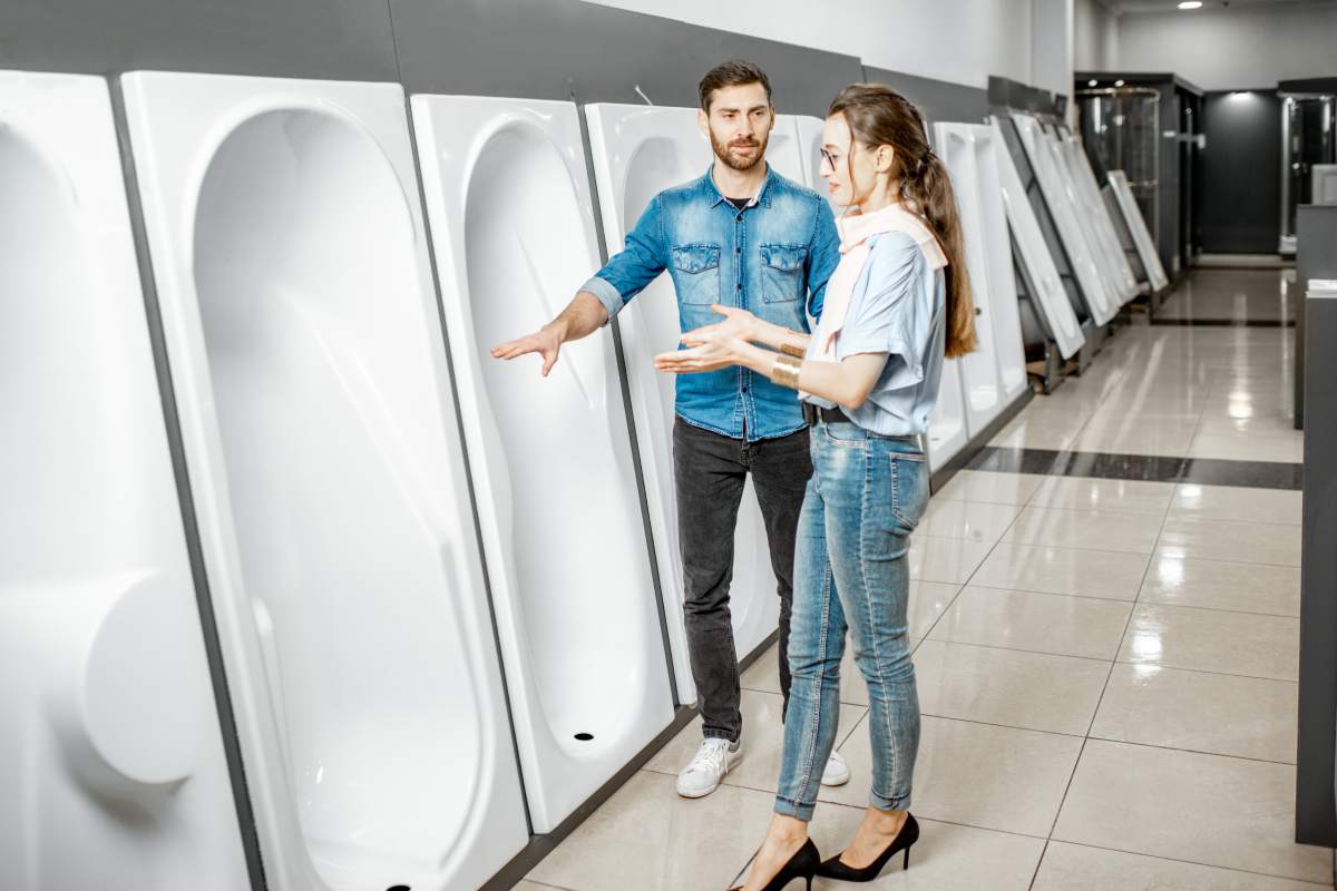 Young couple choosing new bathroom furniture standing near the showcase with white acrylic bathtubs in the plumbing shop