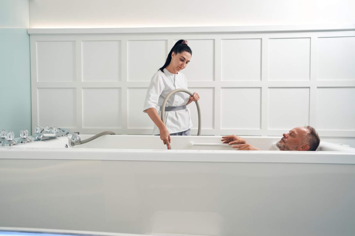 Man relaxing in bathtub while cosmetologist performing hydromassage with special device